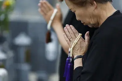 Elderly person praying with beads, hands clasped, in a solemn mood. Another person in background, blurry setting, neutral colors.