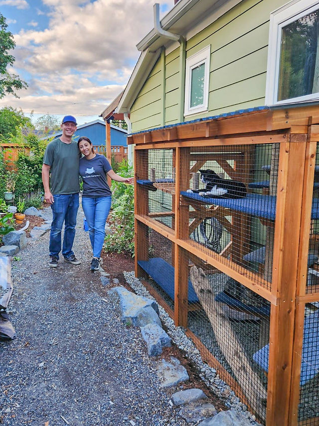 Couple standing next to a custom built wooden catio with multiple levels and a lounging cat, situated beside their home with a landscaped garden path