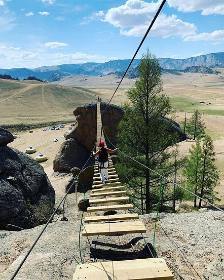 A person wearing a safety harness walks across a wooden slat suspension bridge overlooking a vast rocky valley in Terelj
