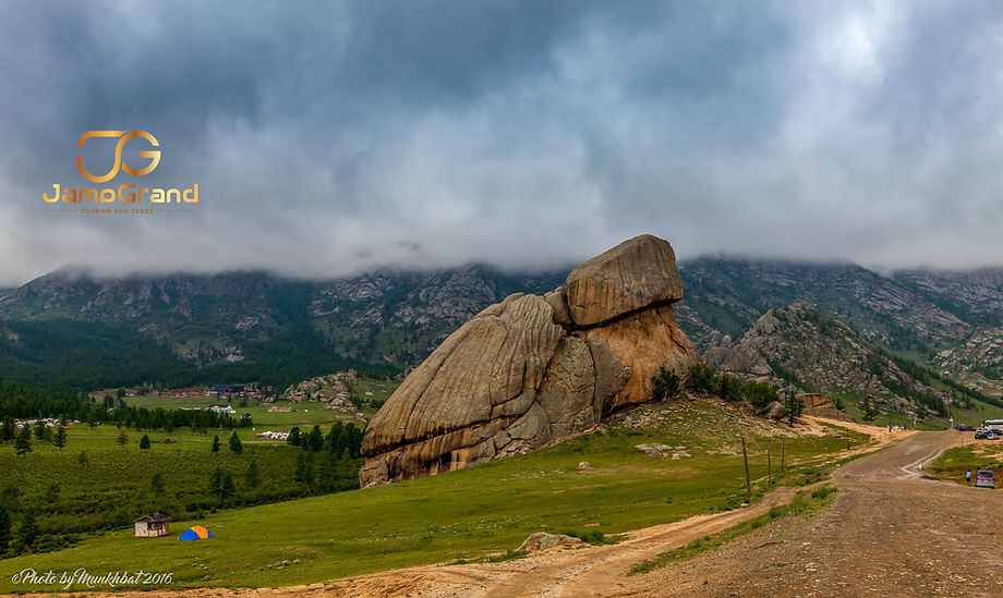 A massive, natural rock formation resembling a giant turtle sitting in a green valley under a cloudy sky