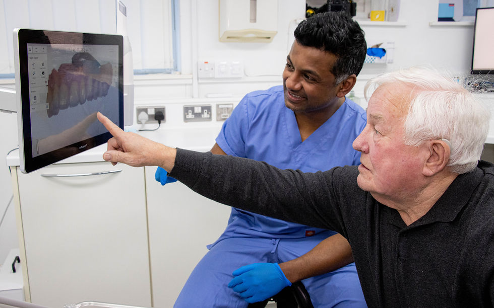 Elderly man points at a dental x-ray on a monitor while a smiling dentist in blue scrubs sits beside him in a clinic.