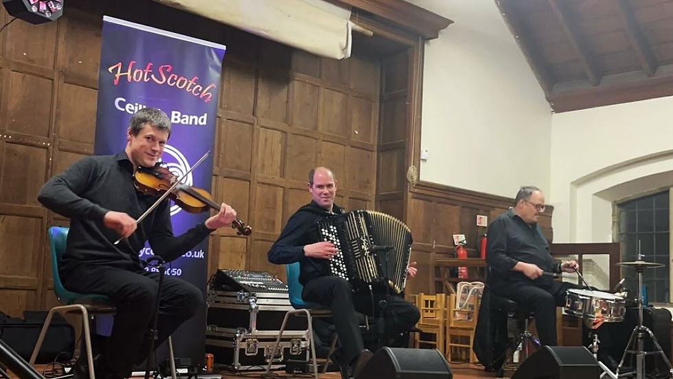 Three musicians perform in a wooden hall. A violinist, accordionist, and drummer sit in front of a "HotScotch Ceilidh Band" banner.