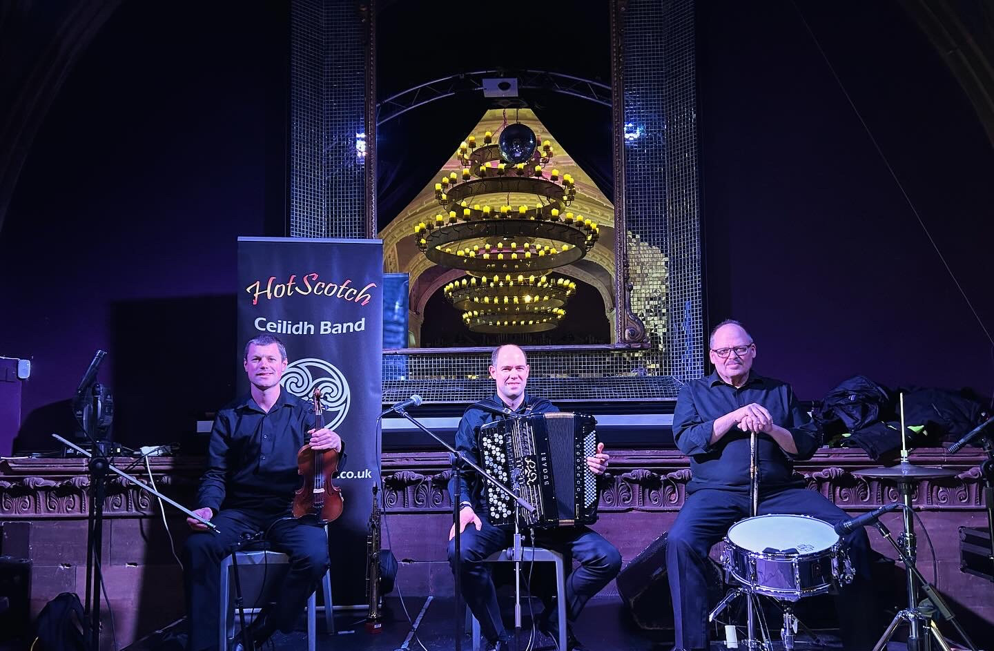 Three-man HotScotch Ceilidh Band onstage with violin, accordion and drum under purple lights, banner behind them.
