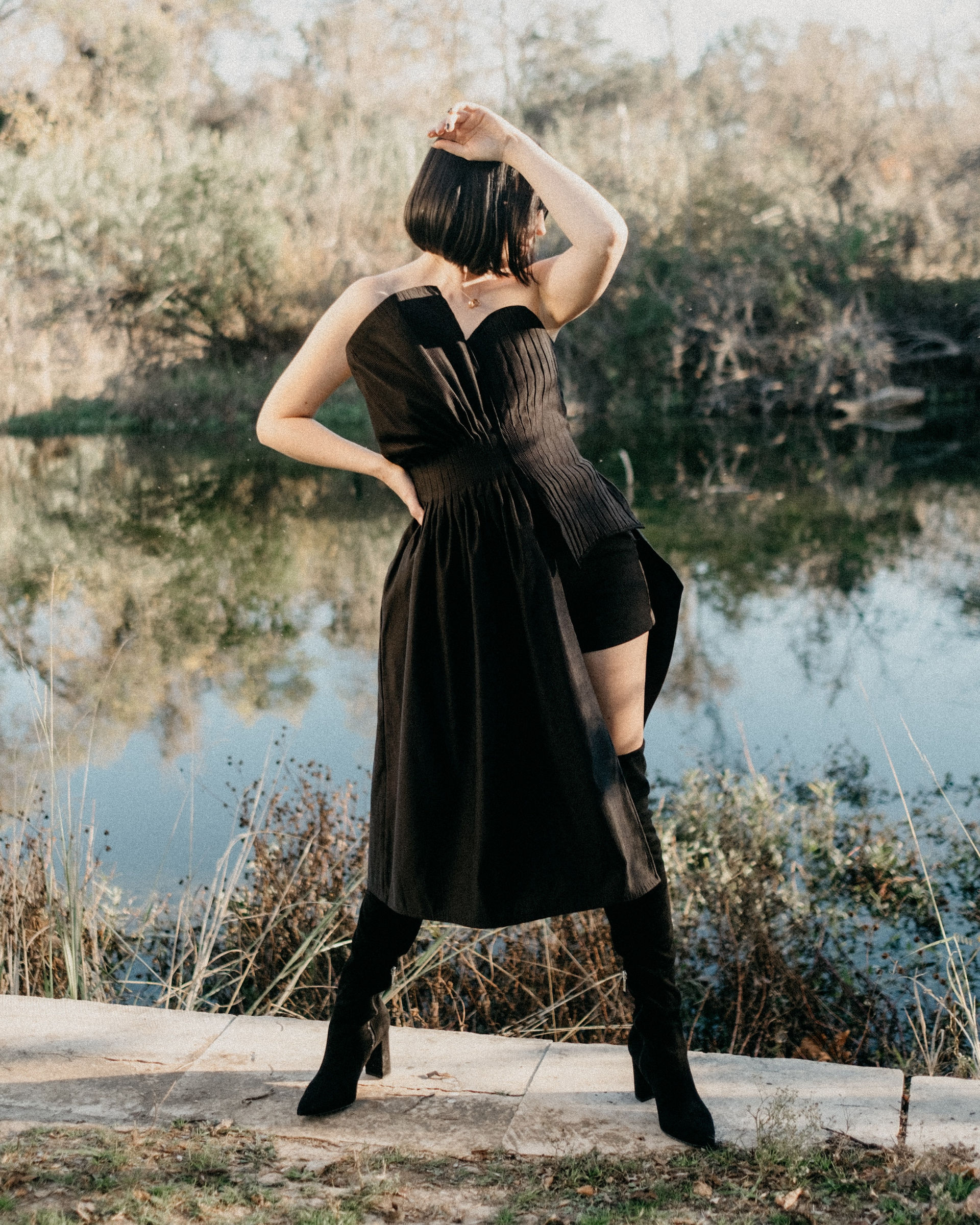 Model standing by the water wearing a black strapless corset top with a pleated half-skirt detail.