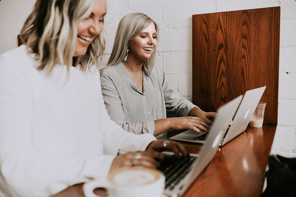 Two blonde women working together on MacBook Pros in a cafe.