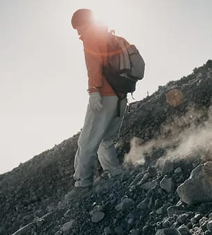 Un escursionista, in controluce e avvolto da una luce soffusa del sole, cammina su un terreno roccioso e scosceso, segno di una scalata impegnativa. Indossa abbigliamento da trekking e uno zaino, simbolo della sua avventura sul vulcano Rinjani