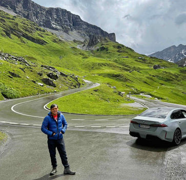 Klausen Pass with a silver car and driver on a wet alpine road amid lush green meadows.