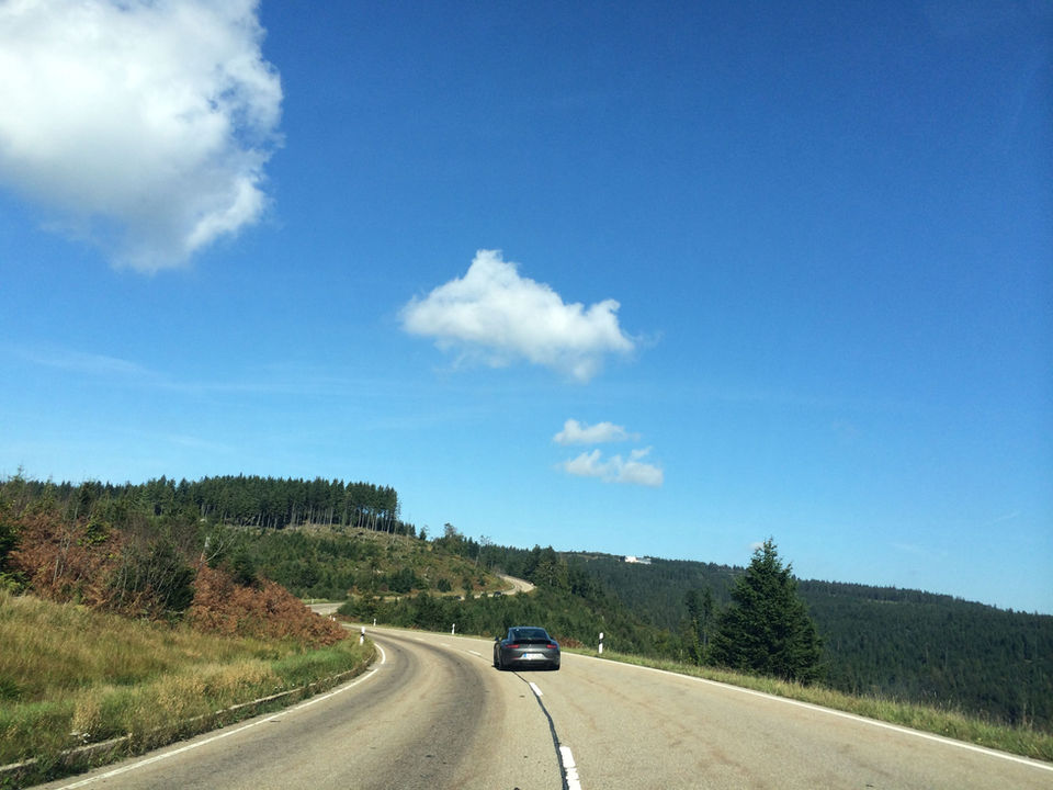 Porsche 911 driving on the Black Forest High Road in Germany, with sweeping curves, forested hills, and open countryside under a clear blue sky.