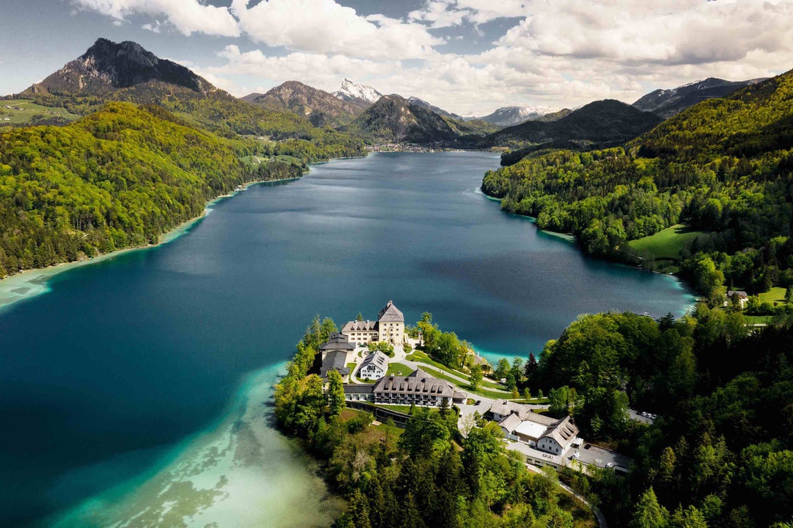 Aerial view of Schloss Fuschl on a wooded peninsula, surrounded by the turquoise waters of Lake Fuschl and alpine mountains in the background.