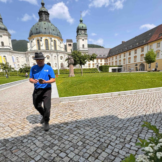 Man in a blue shirt walking across a cobbled courtyard in front of a domed baroque abbey.
