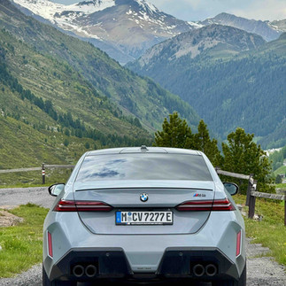 Rear view of a silver BMW sports car parked on a mountain road overlooking an alpine valley.
