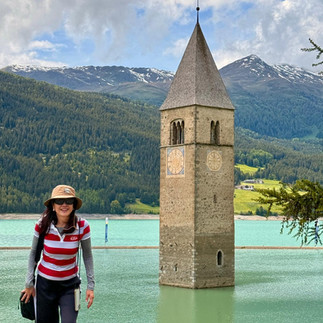 Woman in sunhat and striped top posing in front of the submerged church tower of Reschensee with forested mountains behind.