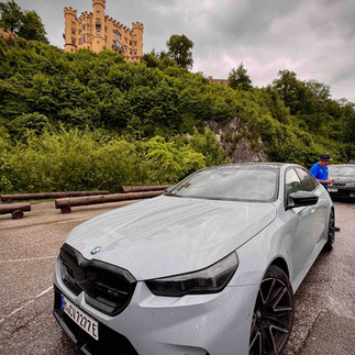 BMW in a car park beneath a yellow hilltop castle surrounded by lush green trees on an overcast day.
