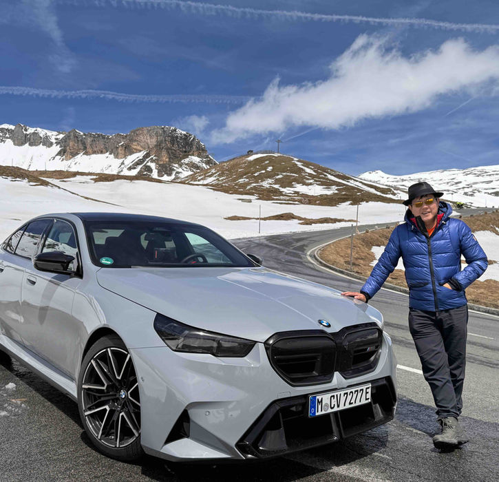 Grey BMW M5 parked on a snowy alpine pass with a smiling guest leaning against the car during a luxury driving tour.