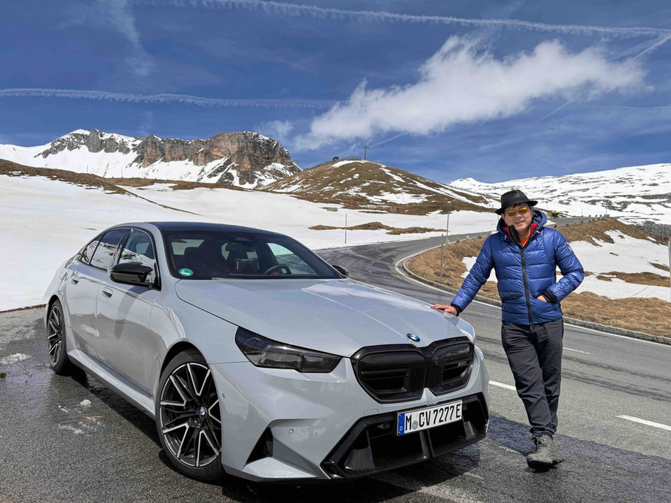 Grey BMW M5 parked on a snowy alpine pass with a smiling guest leaning against the car during a luxury driving tour.