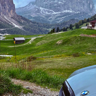 Front of a grey BMW M5 parked beside a green meadow with wooden huts and dramatic Dolomite peaks in the background.