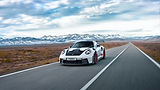 White Porsche sports car with a large rear wing and red wheels driving on an empty straight road, with snow-capped mountains in the distance.