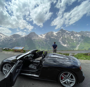Black Audi R8 Spyder parked at a high viewpoint while the driver looks out over rugged, snow-capped alpine peaks.