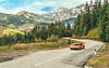 Orange Lamborghini Huracán driving along a winding Alpine mountain road surrounded by forests and dramatic peaks in Europe.