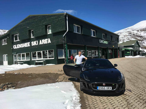 Man leaning against a Jaguar F-Type R at the summit of Scotland’s highest public road pass, snow still on the surrounding peaks.