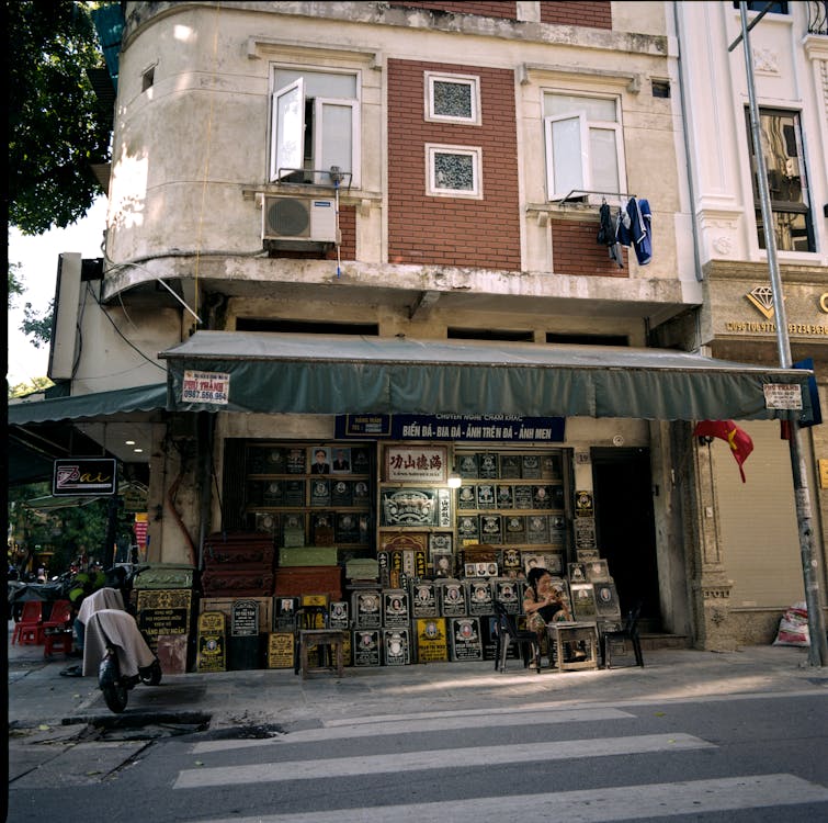 A street-level view of a small storefront featuring a collection of framed artworks or signage displayed outside. The building has a weathered exterior with open windows on the upper floor and an awning covering the shop entrance. A person sits outside on a chair, surrounded by the shop's items, while a motorbike is parked nearby. The scene captures the charm of a bustling, urban neighbourhood.