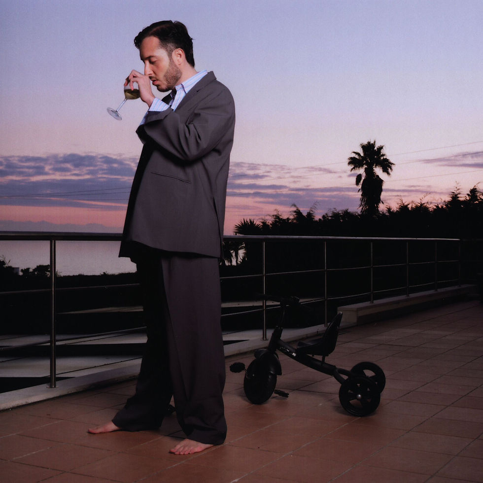 Pedro Mafama retratado em pose contemplativa, de fato largo e descalço, numa varanda ao pôr do sol com um triciclo atrás.