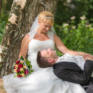 Braut und Bräutigam bei einem Hochzeitsshooting in der Nähe der Waldschänke an der Müritz, wobei die Braut in ihrem weißen Brautkleid an einem Baum sitzt und lächelnd auf den Bräutigam herabschaut, der mit geschlossenen Augen entspannt mit dem Kopf auf ihrem Schoß liegt, während ein rot-orangefarbener Brautstrauß neben ihnen auf dem Kleid liegt und grüne Pflanzen den Hintergrund umgeben.