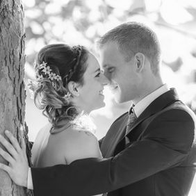 Schwarzweißfoto von Braut und Bräutigam bei einem Hochzeitsshooting an einem Baum beim Schloss Klink, wie sie sich eng gegenüberstehen, sich lächelnd in die Augen sehen und ihre Nasen fast berühren, während der Bräutigam die Braut mit beiden Armen am Baum umrahmt und die Braut mit einer Hand am Baumstamm lehnt.
