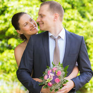 Braut und Bräutigam stehen bei einem Hochzeitsshooting im Schaugarten am Hotel am Tiefwarensee in Waren (Müritz), wobei die Braut den Bräutigam von hinten umarmt, beide sich lächelnd ansehen und der Bräutigam den rosa-lila Brautstrauß vor sich in den Händen hält, während im Hintergrund weiches grünes Laub unscharf verschwimmt.