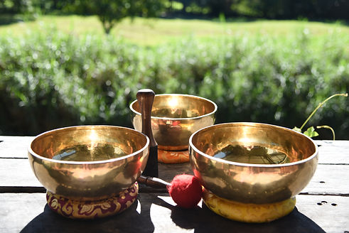 3 gold sound bowls with a red and black mallet, resting on a wooden bench with grass in the background