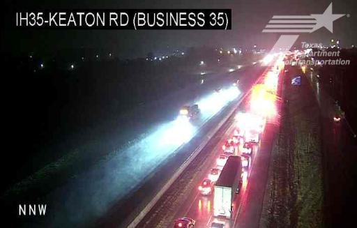 TxDOT traffic camera still labeled IH35 Keaton Rd (Business 35) in Sanger showing emergency lights and backed-up northbound traffic on wet pavement early Thursday