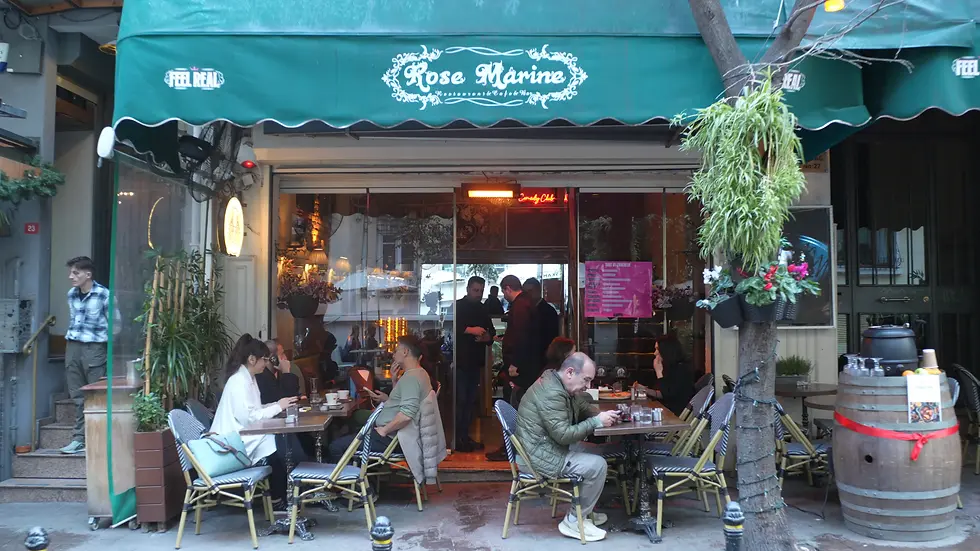 People are sitting in the cafe, chatting under the green awning. There are other customers inside; the sign reads Rose Marine. A warm and friendly atmosphere.