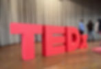 Large, 3D red matte letters "TEDx" installation standing on a wooden stage in front of grey pleated curtains for a TEDx event.