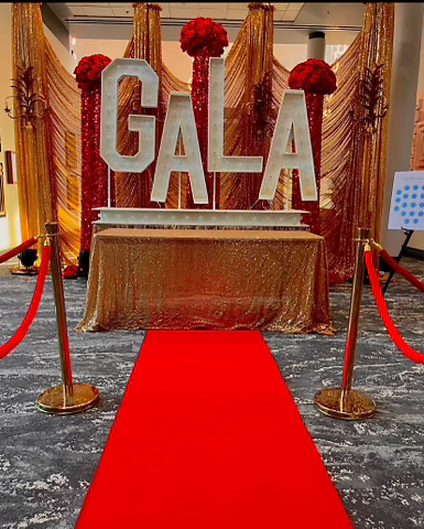 Red carpet entrance to a gala featuring large marquee "GALA" letters, gold sequin table linens, and red velvet stanchion ropes.