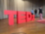 Large, 3D red matte letters "TEDx" installation standing on a wooden stage in front of grey pleated curtains for a TEDx event.