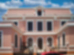 Elegant pink facade of the City Museum of Mérida with arched windows, white trim, and a decorative tiled rooftop, set against a blue sky with scattered clouds.