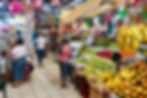 Fruit and grocery section inside Mérida’s San Benito Market with vendors selling bananas, mangos, apples, and grapes, while nearby stalls display household items, bags, and colorful papel picado decorations overhead.