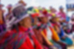 Quechua people in bright, traditional outfits with patterned hats sit outdoors. They are wearing vivid colors and floral decorations on their hats.