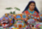 Wixárika woman in colorful traditional attire creating vibrant art with animal motifs.