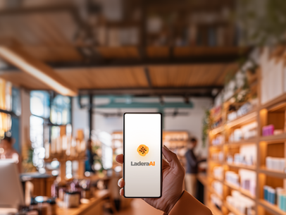 Hand holding a smartphone displaying the Ladera AI logo inside a modern hotel café, with shelves and a service counter softly blurred in the background.