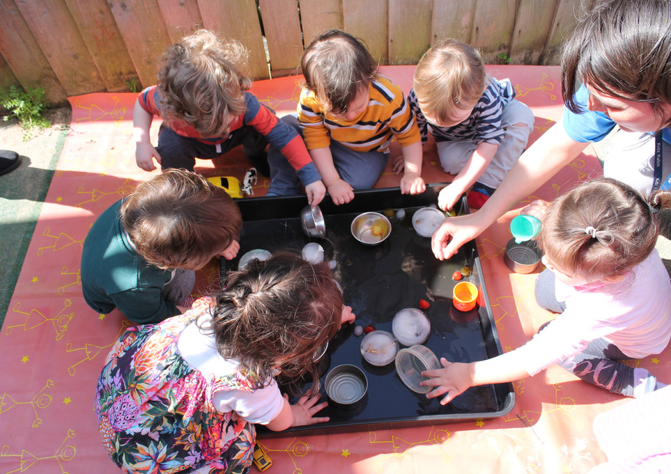 A group of toddlers from Kidz Kabin Nursery in Muswell Hill are sitting outside on a picnic blanket and learning about what happens when ice melts