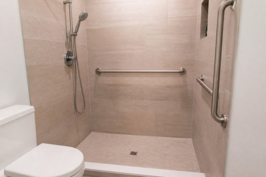 Close-up of bathroom safety features including grab bars installed at proper height, handheld shower wand mounted on wall, and non-slip shower flooring under bright lighting