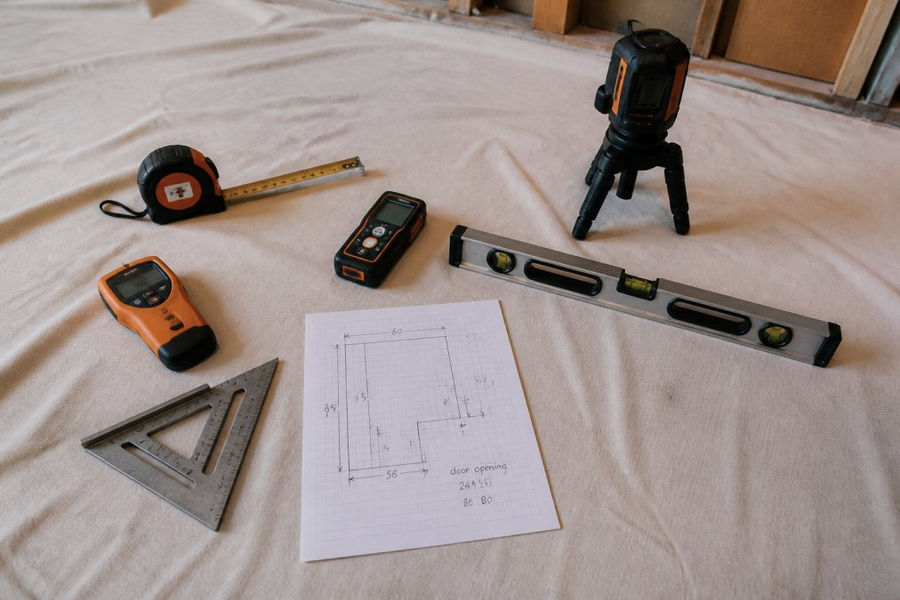 A layout of shower measurement tools on a drop cloth, 25-foot tape measure, laser distance measurer, cross-line laser level on a tripod, 4-foot level, stud finder, speed square, and a printed measurement sheet with handwritten dimensions