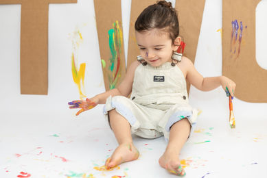 Child smiling with painted hands and feet, sitting in front of colorful mess