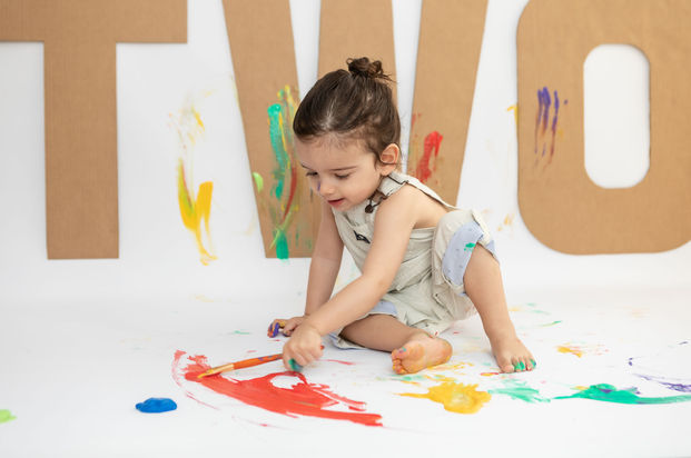 Joyful toddler painting red on floor with big smile, surrounded by splashes of color