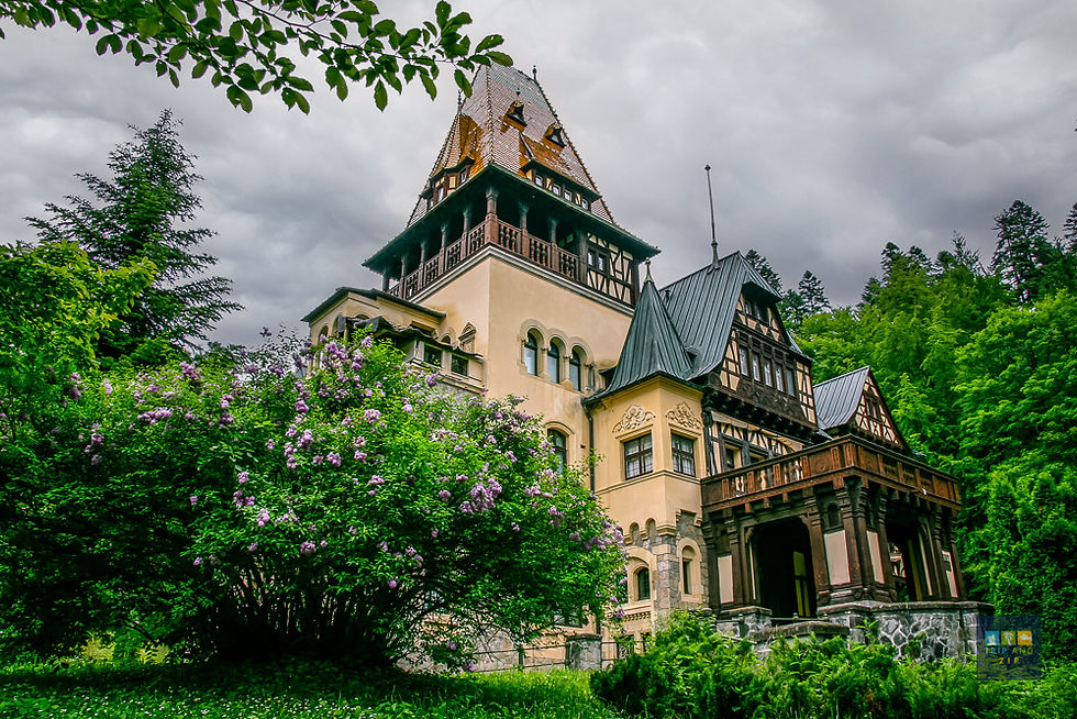 A grand, ornate building amidst lush greenery and purple flowers under a cloudy sky, exuding a serene, historic ambiance.