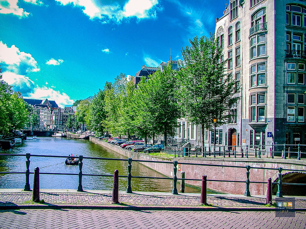 Scenic canal with a small boat in Amsterdam, bordered by trees and historic buildings. Blue sky with clouds adds a calm, vibrant mood.