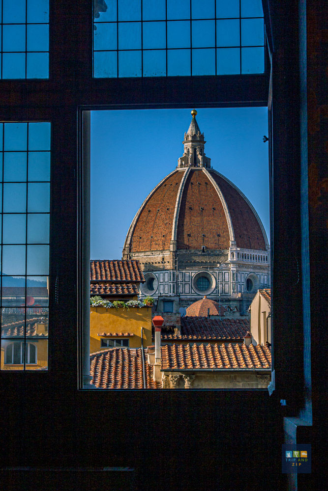 View of Florence's Duomo through a window with grid frames. Clear blue sky, terracotta rooftops, and vibrant yellow buildings.
