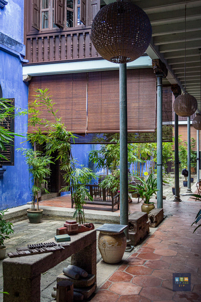 Covered patio with bamboo plants, blue walls, and wooden furniture. Clay pots and stone objects line the walkway. Calm, rustic ambiance.