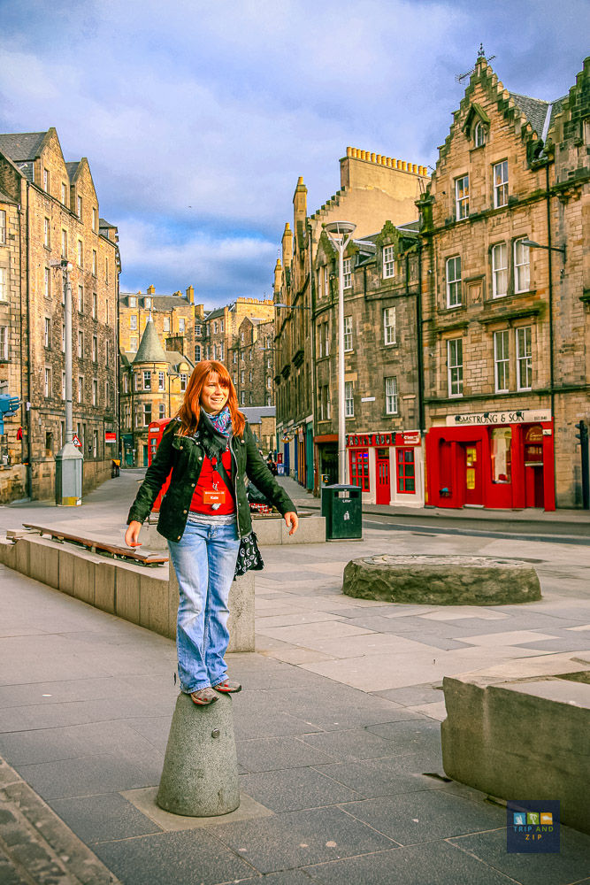 Woman in a red shirt and jeans stands on a post in a cobblestone street. Historic buildings with red doors in the background under a cloudy sky.